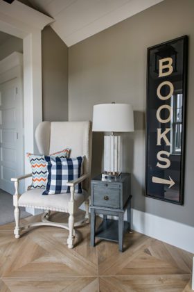 Closeup photo of a while upholstered chair with multi-colored pillows, end table, and a lamp