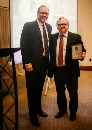 Two men standing and facing the camera, one holding a framed award under his arm