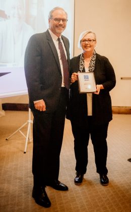 A man standing with a woman who is holding a framed award, both smiling at the camera