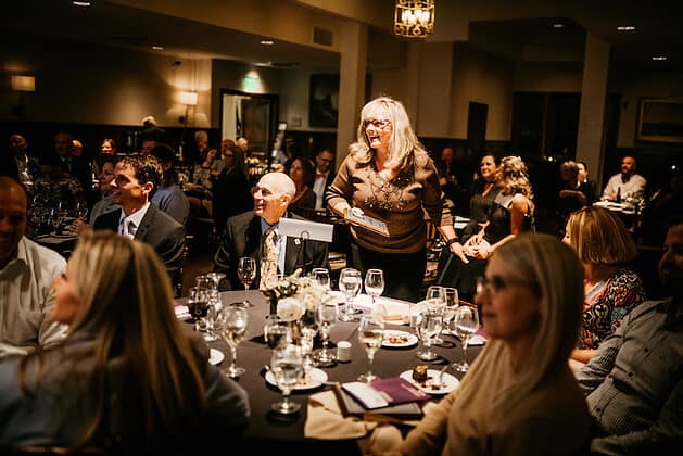 A woman standing and talking with other people seated at dining tables around her