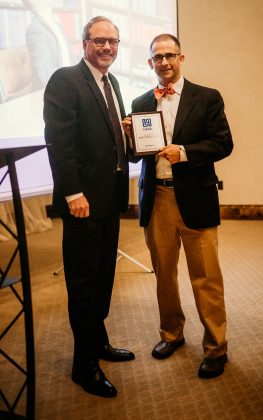 Two men standing and smiling at the camera, one holding a framed award