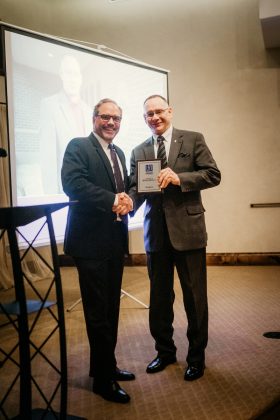 Two men shaking hands and facing the camera, one holding a framed award
