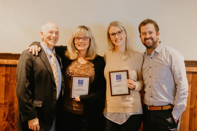A group of four smiling men and women standing and facing the camera, two holding framed awards