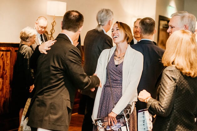 A woman smiling and shaking hands with a man, with other people behind them talking