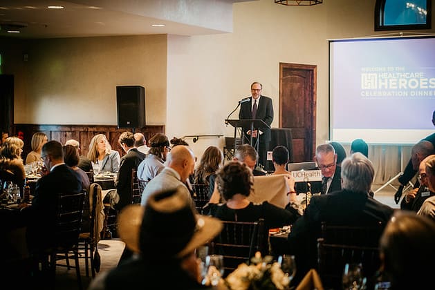 A man standing talking to groups of people seated at dining tables
