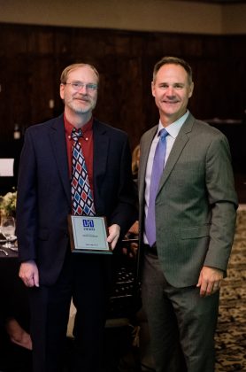 Photo of two men facing the camera, one holding a framed award between them