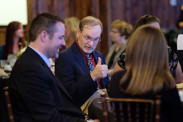 Two men talking, one with a thumbs-up, with other people standing around them
