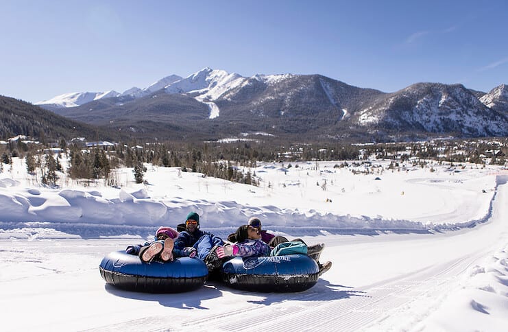 A family of four slides down the Frisco Adventure Park Tubing Hill in Colorado with snow-covered mountains in the background.