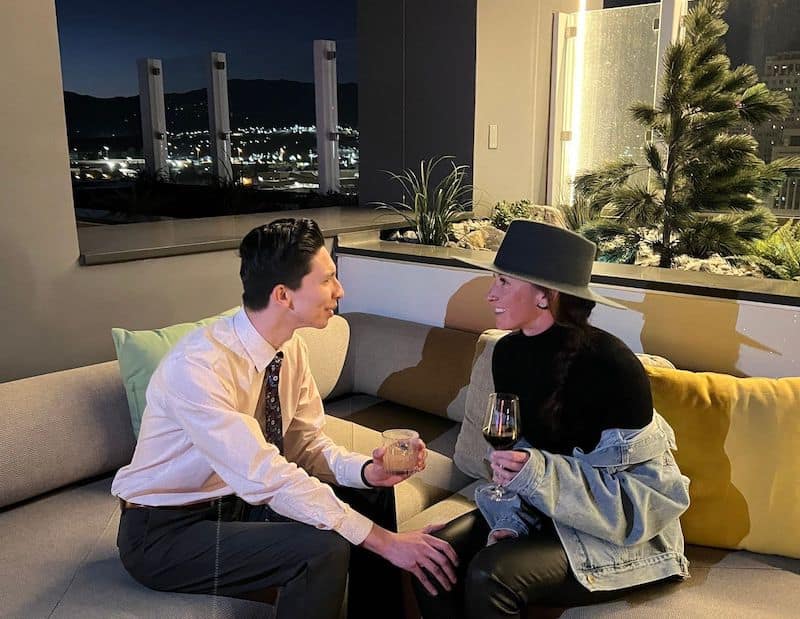 City lights and mountain silhouette line the horizon behind a couple with drinks on the patio at Lumen8 Rooftop Social in Colorado Springs.