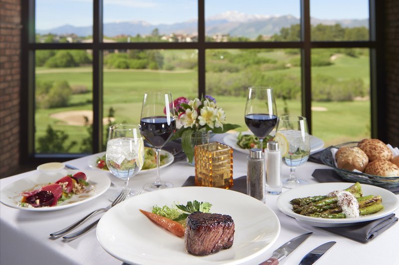 Steak dinner in front of the large windows framing Pikes Peak and golf course views from the Steakhouse at Flying Horse, one of the best restaurants in Colorado Springs. 