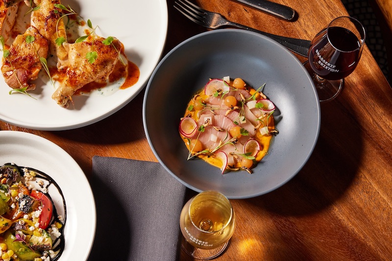 A table full of dishes, including hamachi crude, and wine glasses at Brasserie Brixton, a new Michelin Recommended restaurant in Denver.