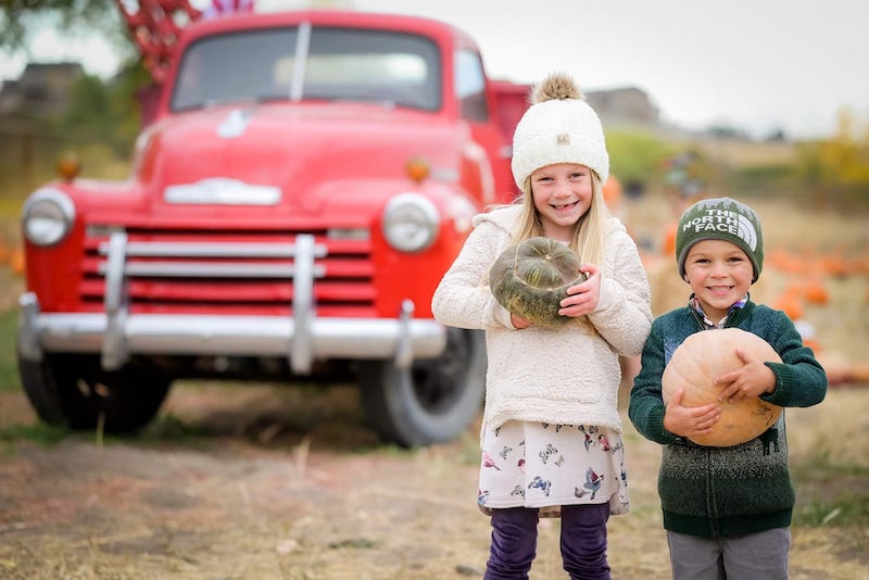 Young boy and girl hold pumpkins and stand in front of old red pickup truck at Gilly's Pumpkin Patch in Colorado