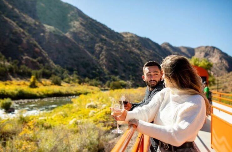 A man and woman hold wine glasses on open car of Royal Gorge Railroad, passing by Colorado fall colors