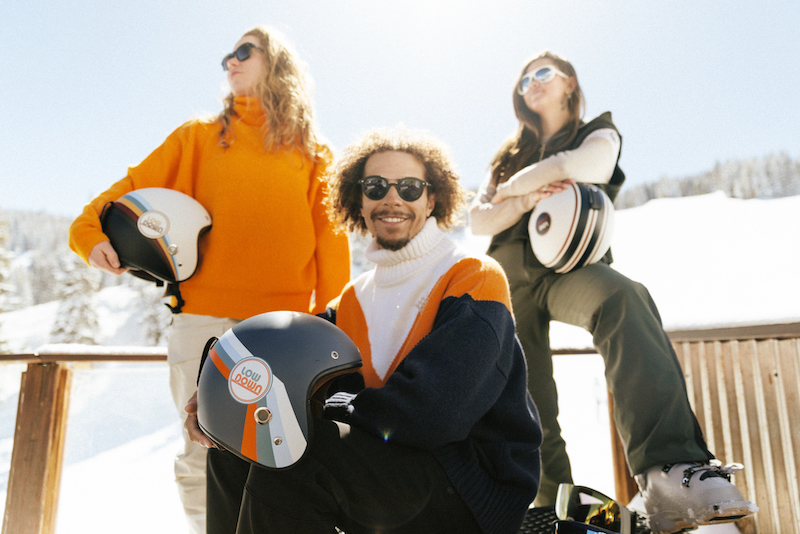Skiers and snowboarders hold their LowDown helmets in front of ski slopes.