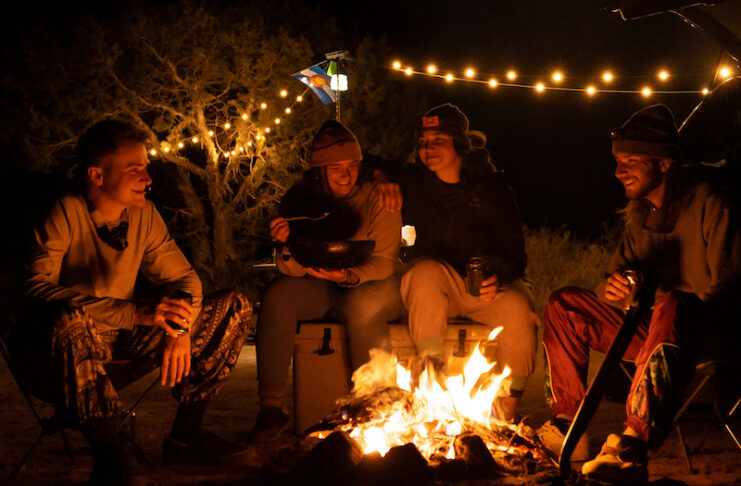 A group of campers sits around a campfire with string lights hanging overhead