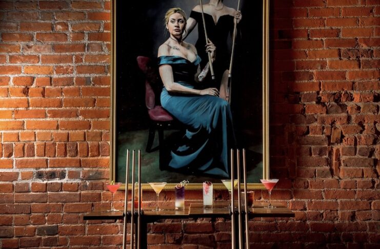 Pool cues and cocktails in front of a painted portrait of women in formal gowns with pool cues