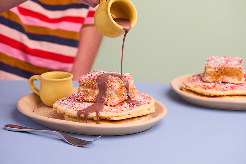 An person in a bright, striped shirt pours chocolate syrup on Snooze pancakes sprinkled with peppermint crumbles