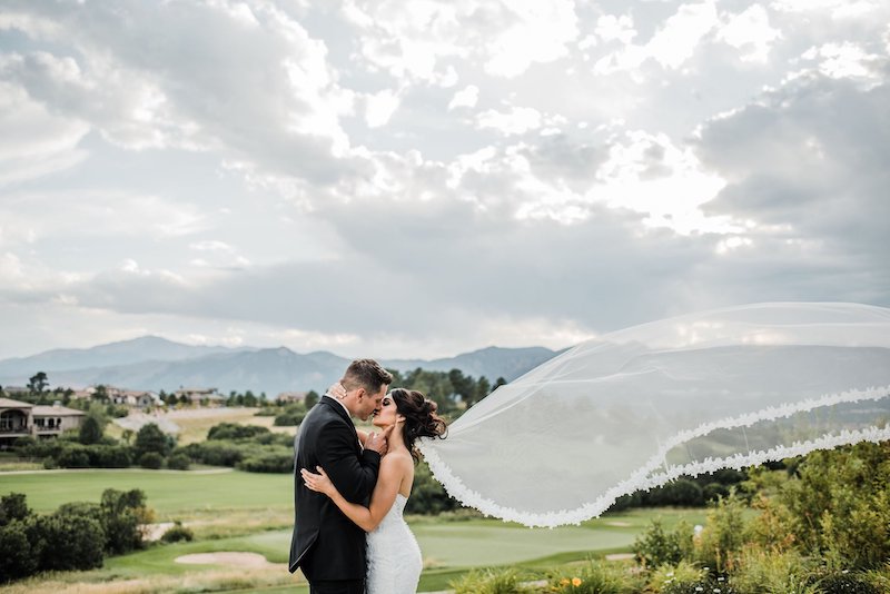 A bride's veil flutters in the wind as the bride and groom kiss. Golf course and Pikes Peak views like this make Flying Horse one of the most scenic wedding venues in Colorado Springs.