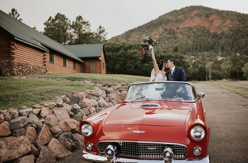 A bride and groom kiss in a red classic sports car before leaving their wedding at Pinecrest Weddings venue in Palmer Lake.