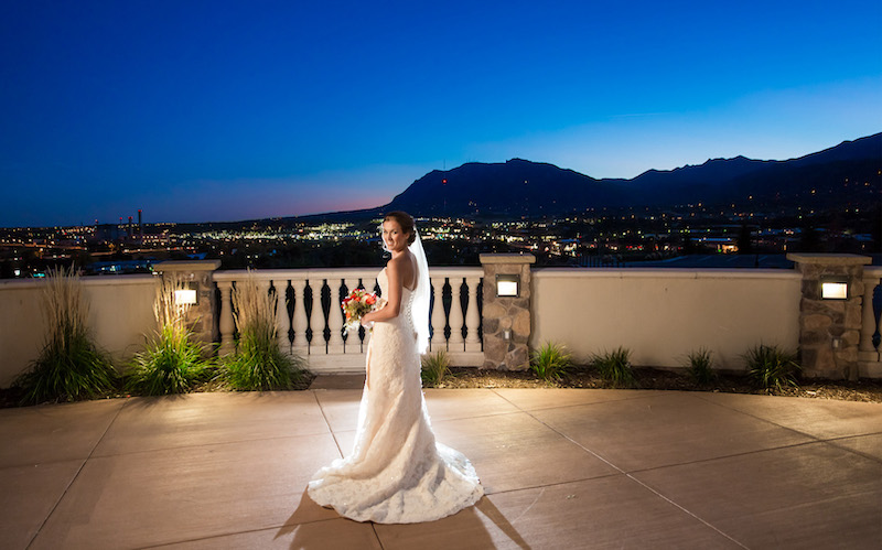 A bride at the Pinery at the Hill overlooks Colorado Springs as city lights and Cheyenne Mountain glow at sunset.