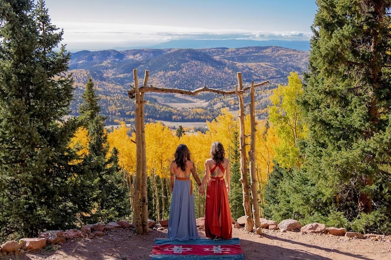 Two brides strand in front of the valley overlook filled with golden aspen trees at Rhize Weddings, one of the most scenic wedding venues in Colorado Springs. 