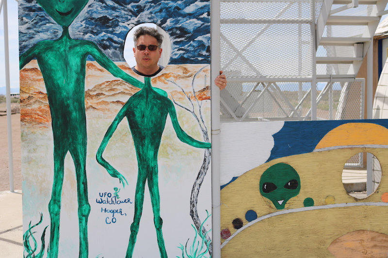 Steven Hayward shows his face through a photo cutout of aliens at the UFO Watchtower in Hooper, Colorado. He explores the misadventures of living in Colorado Springs in his humor articles for Springs magazine.