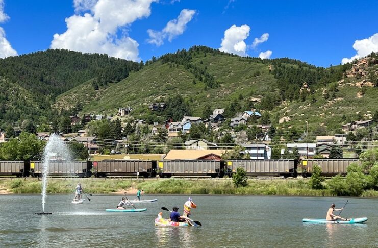 People paddle SUPs and an inflatable swan on Palmer Lake as a train passes by. Mountains line the horizon. It all makes Palmer Lake one of the best lakes in Colorado Springs.