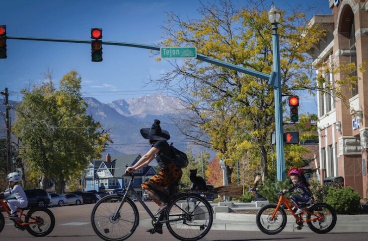 A mom witch and kids in Halloween costumes ride bikes across the street with Pikes Peak in the background on the Witches, Warlocks and Ghouls Ride, a popular fundraising ride for Halloween in Colorado Springs.