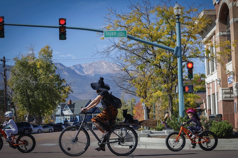A mom witch and kids in Halloween costumes ride bikes across the street with Pikes Peak in the background on the Witches, Warlocks and Ghouls Ride, a popular fundraising ride for Halloween in Colorado Springs. 