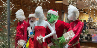 A holiday storefront display featuring two people dressed as Santa and the Grinch posing among three mannequins in red sequin dresses and white winter hats. The mannequins hold wrapped gifts, and the scene is surrounded by evergreen branches and warm string lights reflecting off the shop’s windows.