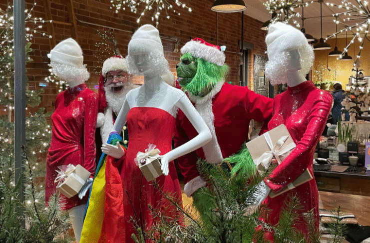 A holiday storefront display featuring two people dressed as Santa and the Grinch posing among three mannequins in red sequin dresses and white winter hats. The mannequins hold wrapped gifts, and the scene is surrounded by evergreen branches and warm string lights reflecting off the shop’s windows.