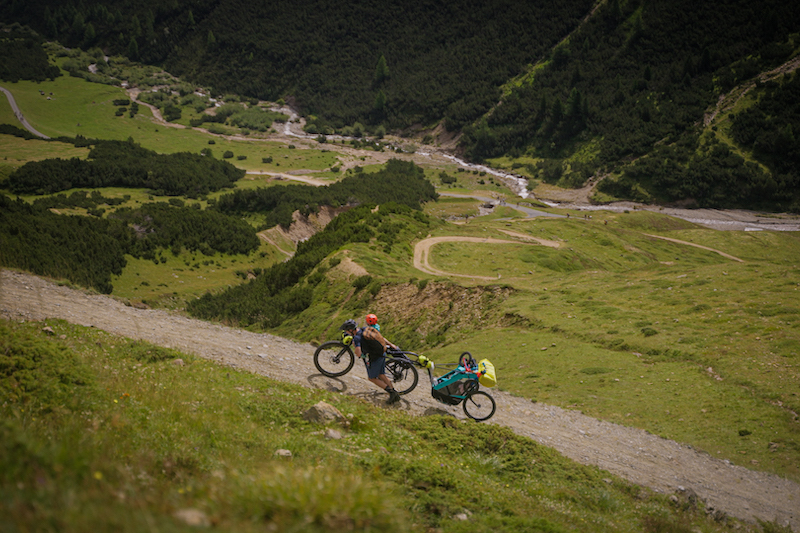 A father pushes his bike and bike trailer carrying his toddler up a steep mountain road in the film Emil and Karl - Little Wheels Big Mountains on the Banff Film Festival World Tour.