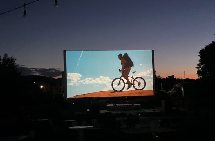 An outdoor movie screen shows a film at sunset for the CO150 Film Festival