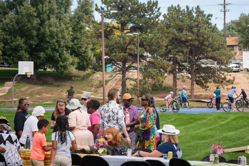 Neighbors talk around a long table in the park while kids ride bikes at the Hey Neighbor Hillside Community Lunch. 