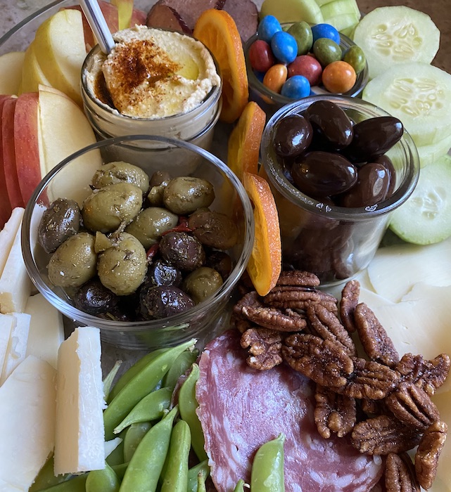 A charcuterie board with hummus, olives, cucumbers, snap peas, walnuts and more.