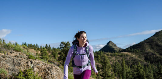 Our Favorite Hiking Gear Now A woman hikes in the mountains wearing an Icebreaker Quantum Hoodie, one of our favorite hiking gear picks.